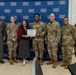 A group of seven individuals pose in front of a USO backdrop, with two people in the center holding a certificate or award document. Six of the individuals are wearing military camouflage (OCP) uniforms or a flight suit, while one civilian woman in a red jacket and black skirt stands at the center. This appears to be a formal recognition or award ceremony at a USO location.