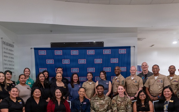A large group of approximately 30 individuals poses for a formal group photograph in front of a USO branded step-and-repeat backdrop. The group includes both military personnel in various uniforms (Army combat uniforms, Navy/Marine dress uniforms, and flight suits) and civilians in business attire. The setting appears to be inside a USO facility, with the USO mission statement visible on the wall to the left.