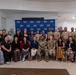 A large group of approximately 30 individuals poses for a formal group photograph in front of a USO branded step-and-repeat backdrop. The group includes both military personnel in various uniforms (Army combat uniforms, Navy/Marine dress uniforms, and flight suits) and civilians in business attire. The setting appears to be inside a USO facility, with the USO mission statement visible on the wall to the left.