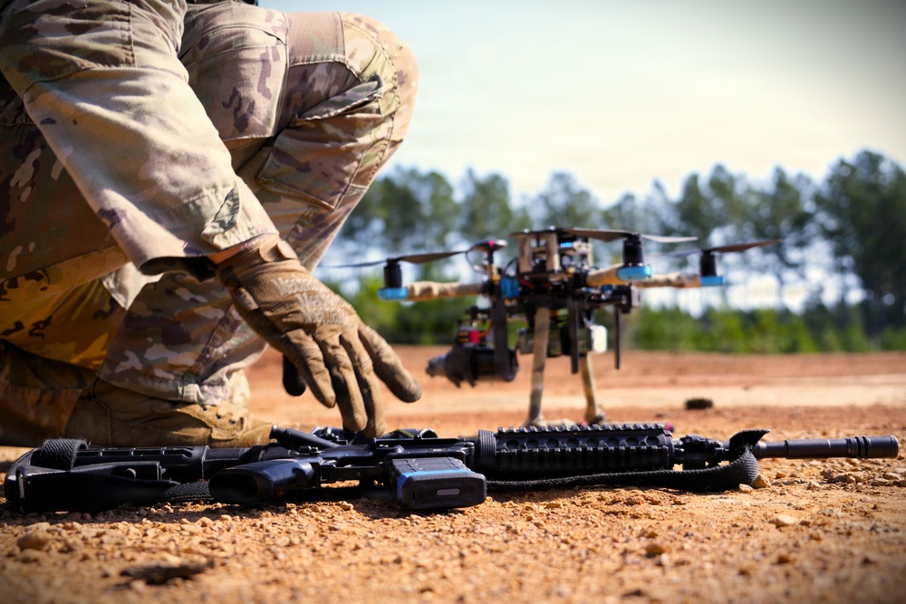 A military or law enforcement personnel in MultiCam/OCP camouflage uniform is crouching near equipment on a dirt surface. In the foreground, an assault rifle with rail accessories and a radio/communication device are laid on the ground. A multi-rotor drone (quadcopter or hexacopter) with a camera gimbal is visible in the background.