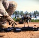 A military or law enforcement personnel in MultiCam/OCP camouflage uniform is crouching near equipment on a dirt surface. In the foreground, an assault rifle with rail accessories and a radio/communication device are laid on the ground. A multi-rotor drone (quadcopter or hexacopter) with a camera gimbal is visible in the background.