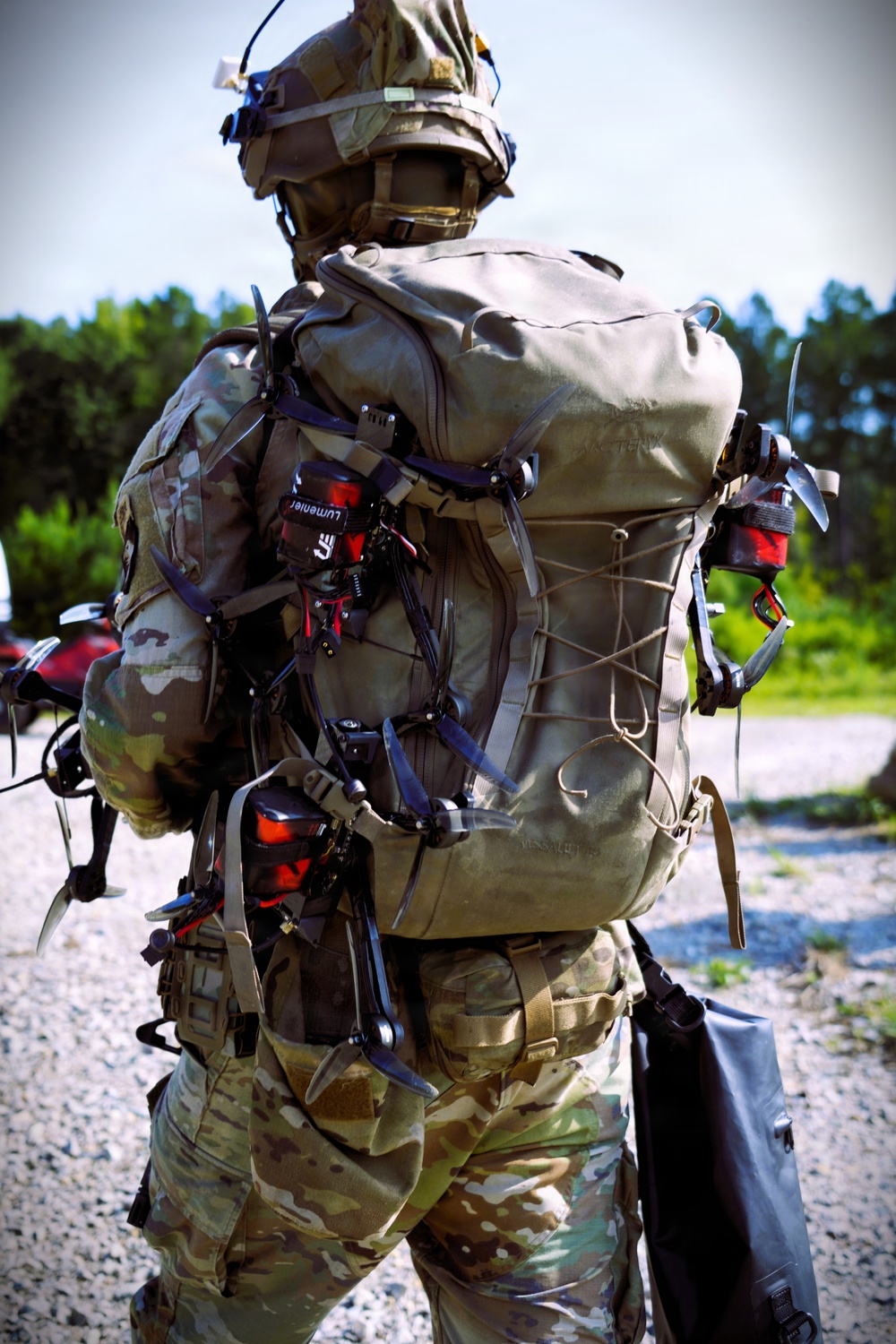 A military soldier photographed from behind is carrying a large tactical rucksack with multiple small drones (FPV-style quadcopters) strapped to the exterior. The soldier is wearing full multicam camouflage uniform, helmet with communications equipment, and appears to be in a training or staging area with gravel ground and tree line in the background. The drones are secured to the pack using straps and bungee cords, with visible red motor housings and propellers.