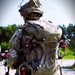 A military soldier photographed from behind is carrying a large tactical rucksack with multiple small drones (FPV-style quadcopters) strapped to the exterior. The soldier is wearing full multicam camouflage uniform, helmet with communications equipment, and appears to be in a training or staging area with gravel ground and tree line in the background. The drones are secured to the pack using straps and bungee cords, with visible red motor housings and propellers.