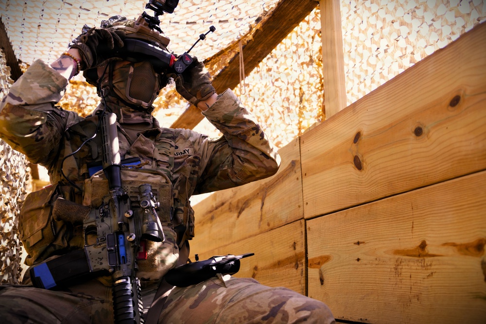 A U.S. Army soldier in full tactical gear is positioned behind a wooden barrier under camouflage netting. The soldier is adjusting head-mounted equipment, possibly night-vision or augmented reality goggles, while carrying an assault rifle. The setting appears to be a forward operating base or training environment.