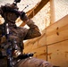 A U.S. Army soldier in full tactical gear is positioned behind a wooden barrier under camouflage netting. The soldier is adjusting head-mounted equipment, possibly night-vision or augmented reality goggles, while carrying an assault rifle. The setting appears to be a forward operating base or training environment.