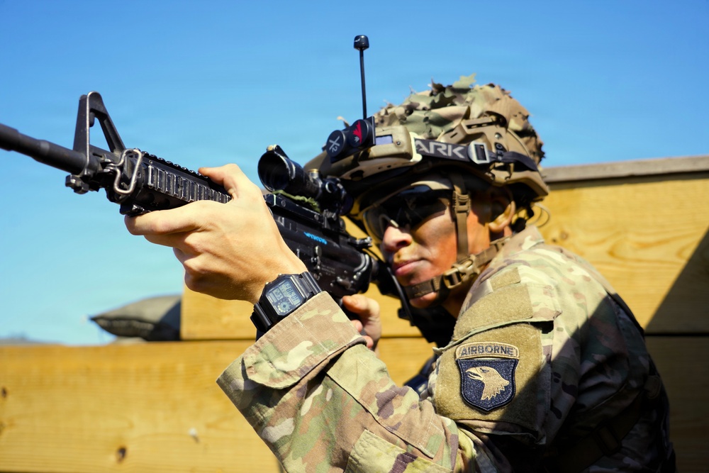 A U.S. Army soldier wearing MultiCam (OCP) camouflage and a tactical helmet is holding an M4-style carbine in a ready/aiming position. The soldier displays a 101st Airborne Division patch on the left shoulder. The image appears to be taken during a training exercise or operational deployment.