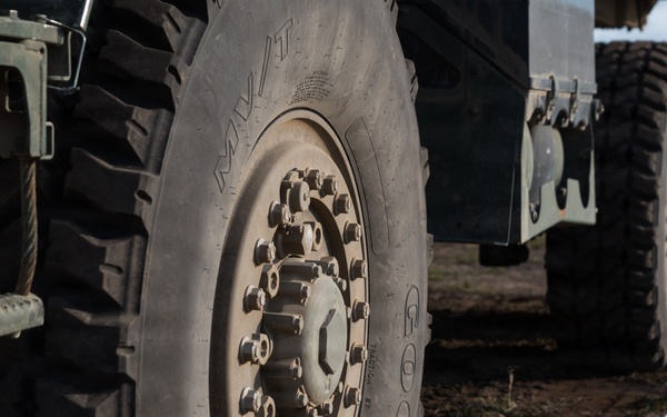 A close-up ground-level photograph of a large military wheeled vehicle, showing multiple heavy-duty off-road tires in perspective. The tires are branded 'Goodyear' with an M/T (mud terrain) tread pattern and feature large multi-bolt hub assemblies. The vehicle is parked on a dirt field with a boundary rope and marker stake visible in the foreground.