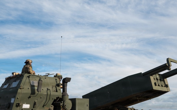 The image shows a military multiple launch rocket system (MLRS) mounted on a heavy tactical wheeled vehicle, consistent with an M142 HIMARS or M270 MLRS platform. The launcher pod is elevated to a firing position in an open field environment. A single soldier is visible in the operator's position atop the cab.