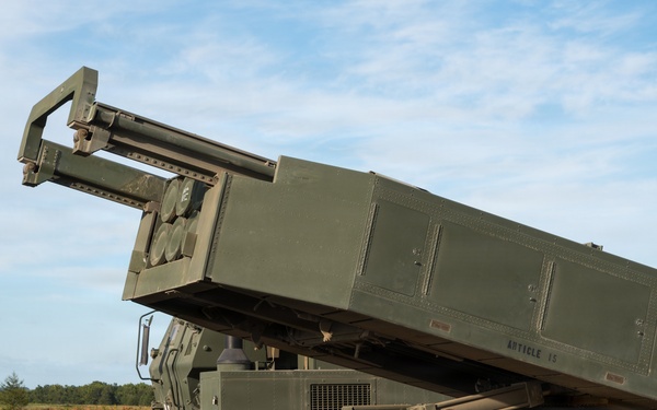 The image shows a military multiple launch rocket system (MLRS) mounted on a wheeled tactical vehicle, with the launcher pod elevated at an angle. The system is positioned on open, unpaved terrain with a rural landscape visible in the background. The vehicle appears to be in a static display or pre-firing position.