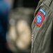 The image shows a close-up of a military uniform sleeve featuring the distinctive patch of the 82nd Airborne Division, United States Army. The patch consists of a red square background with a blue circle containing the letters 'AA' (All American), topped by a blue 'AIRBORNE' tab. A secondary insignia, partially visible below, appears to be a glider or parachutist badge.