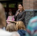 A woman in a gray blazer is speaking at a podium while holding a microphone at what appears to be an outdoor public event or ceremony. She is positioned in front of a brick building with a commemorative or informational plaque mounted on the wall. An audience is visible in the foreground, with additional attendees standing behind the speaker.