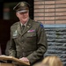 A senior U.S. Army officer in dress green uniform is speaking at a podium, likely at a memorial or commemorative ceremony. He wears multiple service ribbons, unit insignia including what appears to be an airborne patch on his left shoulder, and a peaked officer's cap with the U.S. Army eagle emblem. The setting appears to be an outdoor or semi-covered venue with a brick structure and an engraved memorial plaque visible in the background.