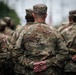 The image shows a group of military personnel in formation, all wearing matching camouflage uniforms consistent with the US Army OCP (Operational Camouflage Pattern). The central figure, whose name tape reads 'BALL,' stands with hands clasped behind their back in a parade rest or at-ease posture. The photo is taken from behind, with shallow depth of field blurring the background.