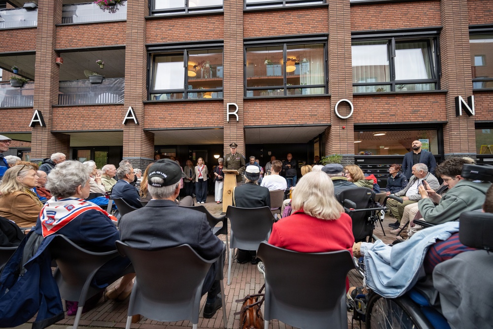 An outdoor public gathering or ceremony is taking place in front of a brick building with the partial name 'AARON' visible on its facade. A uniformed military officer stands at a podium addressing a seated audience of predominantly older adults. The event appears to be a formal commemoration or memorial ceremony, with some attendees wearing Norwegian national scarves.