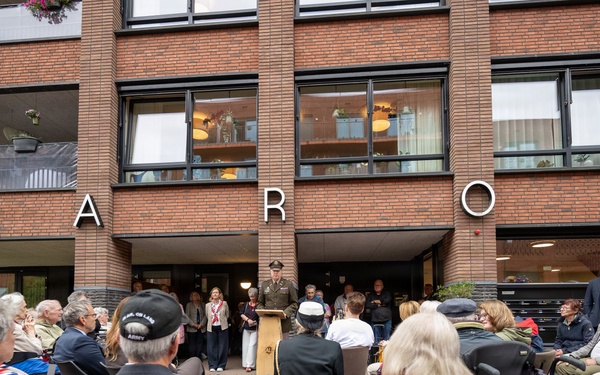 An outdoor public gathering or ceremony is taking place in front of a brick building with the partial name 'AARON' visible on its facade. A uniformed military officer stands at a podium addressing a seated audience of predominantly older adults. The event appears to be a formal commemoration or memorial ceremony, with some attendees wearing Norwegian national scarves.