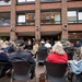 An outdoor public gathering or ceremony is taking place in front of a brick building with the partial name 'AARON' visible on its facade. A uniformed military officer stands at a podium addressing a seated audience of predominantly older adults. The event appears to be a formal commemoration or memorial ceremony, with some attendees wearing Norwegian national scarves.