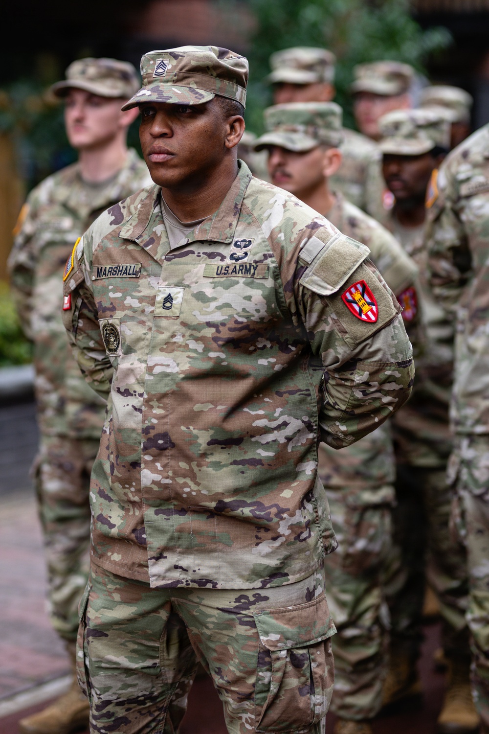 A U.S. Army Staff Sergeant named Marshall stands at parade rest in the foreground, wearing the Army Combat Uniform (ACU) in OCP (Operational Camouflage Pattern). He displays multiple insignia including airborne wings, a unit patch on the right shoulder, and what appears to be a Corps of Engineers patch. Several other soldiers in similar uniforms are visible in the background, suggesting a formation or military ceremony.