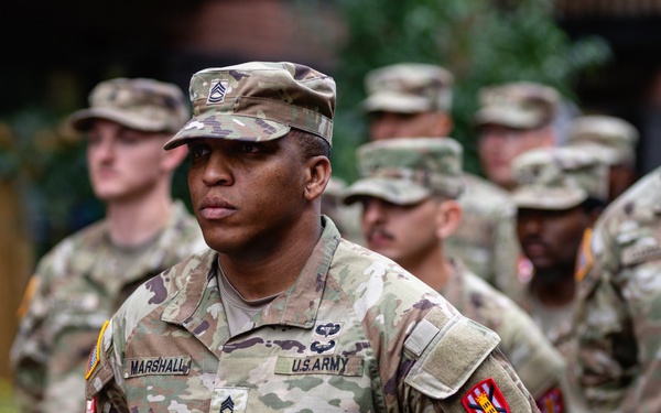 A U.S. Army Staff Sergeant named Marshall stands at parade rest in the foreground, wearing the Army Combat Uniform (ACU) in OCP (Operational Camouflage Pattern). He displays multiple insignia including airborne wings, a unit patch on the right shoulder, and what appears to be a Corps of Engineers patch. Several other soldiers in similar uniforms are visible in the background, suggesting a formation or military ceremony.