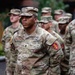 A U.S. Army Staff Sergeant named Marshall stands at parade rest in the foreground, wearing the Army Combat Uniform (ACU) in OCP (Operational Camouflage Pattern). He displays multiple insignia including airborne wings, a unit patch on the right shoulder, and what appears to be a Corps of Engineers patch. Several other soldiers in similar uniforms are visible in the background, suggesting a formation or military ceremony.