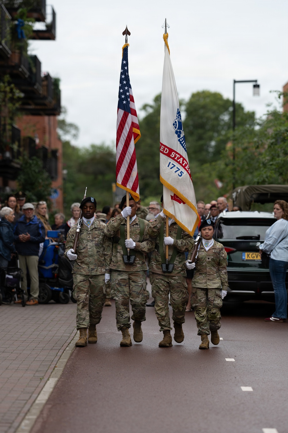 A U.S. Army color guard unit marches in a ceremonial parade along a brick-paved street, carrying the American flag and a United States Army flag dated 1775. The soldiers are dressed in MultiCam camouflage uniforms with black berets and white gloves, flanked by rifle bearers. Civilian spectators line the sidewalk, and the setting appears to be a European town based on the architecture and a visible license plate reading 'KGV'.