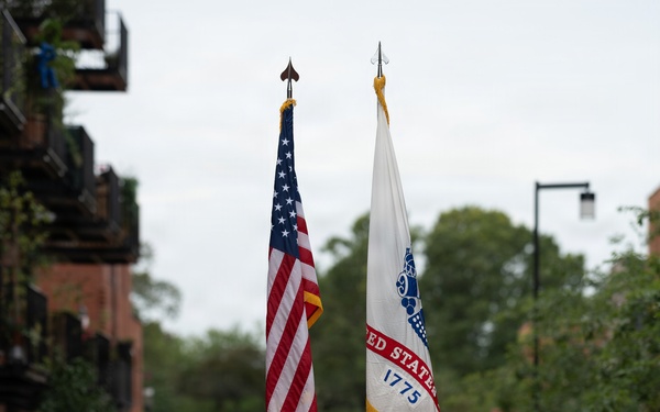 A U.S. Army color guard unit marches in a ceremonial parade along a brick-paved street, carrying the American flag and a United States Army flag dated 1775. The soldiers are dressed in MultiCam camouflage uniforms with black berets and white gloves, flanked by rifle bearers. Civilian spectators line the sidewalk, and the setting appears to be a European town based on the architecture and a visible license plate reading 'KGV'.
