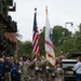 A U.S. Army color guard unit marches in a ceremonial parade along a brick-paved street, carrying the American flag and a United States Army flag dated 1775. The soldiers are dressed in MultiCam camouflage uniforms with black berets and white gloves, flanked by rifle bearers. Civilian spectators line the sidewalk, and the setting appears to be a European town based on the architecture and a visible license plate reading 'KGV'.