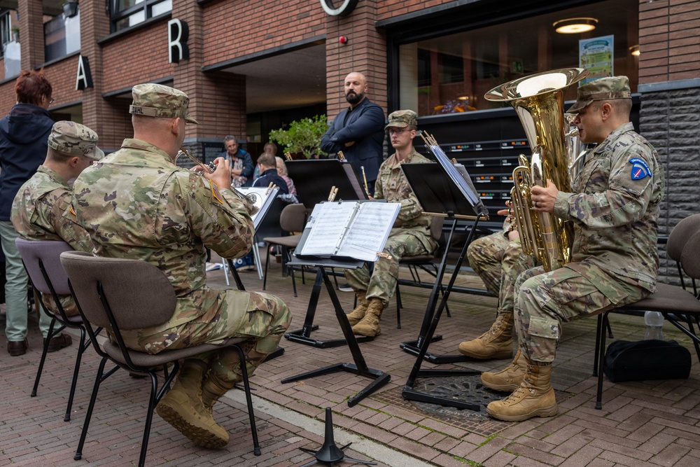 A group of U.S. Army soldiers in MultiCam camouflage uniforms are performing as a brass ensemble in an outdoor public space, likely a European town square or shopping area. The musicians are seated with music stands holding sheet music, playing instruments including a tuba and trumpets. Civilian onlookers are visible in the background observing the performance.