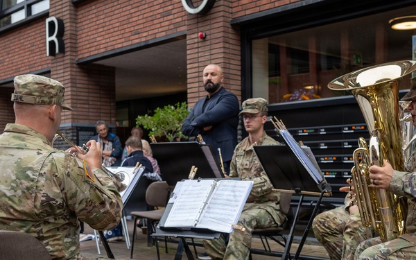 A group of U.S. Army soldiers in MultiCam camouflage uniforms are performing as a brass ensemble in an outdoor public space, likely a European town square or shopping area. The musicians are seated with music stands holding sheet music, playing instruments including a tuba and trumpets. Civilian onlookers are visible in the background observing the performance.