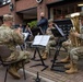 A group of U.S. Army soldiers in MultiCam camouflage uniforms are performing as a brass ensemble in an outdoor public space, likely a European town square or shopping area. The musicians are seated with music stands holding sheet music, playing instruments including a tuba and trumpets. Civilian onlookers are visible in the background observing the performance.