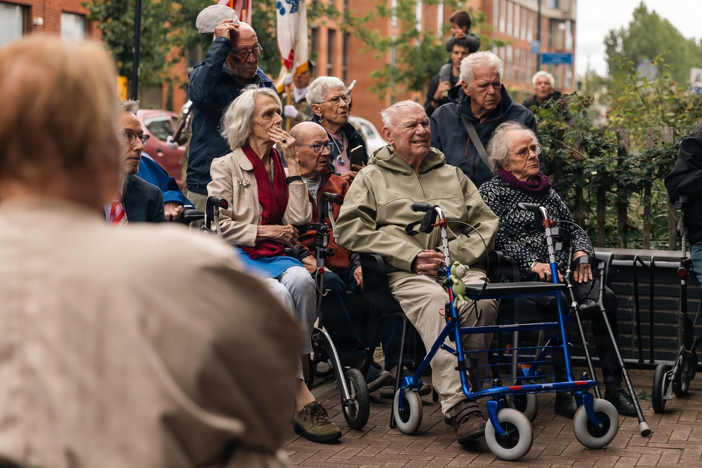 A group of elderly individuals gathered outdoors in an urban setting, many using mobility aids such as wheelchairs and rollators. They appear to be watching or attending a public event or ceremony, suggested by the presence of flags in the background. The scene takes place on a brick-paved street with residential or institutional buildings visible behind them.