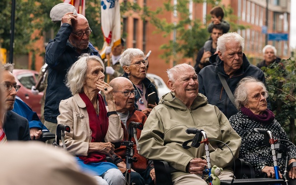 A group of elderly individuals gathered outdoors in an urban setting, many using mobility aids such as wheelchairs and rollators. They appear to be watching or attending a public event or ceremony, suggested by the presence of flags in the background. The scene takes place on a brick-paved street with residential or institutional buildings visible behind them.
