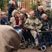 A group of elderly individuals gathered outdoors in an urban setting, many using mobility aids such as wheelchairs and rollators. They appear to be watching or attending a public event or ceremony, suggested by the presence of flags in the background. The scene takes place on a brick-paved street with residential or institutional buildings visible behind them.