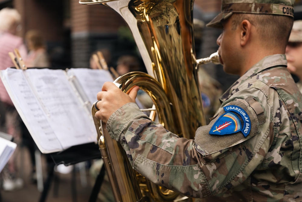 A U.S. Army soldier assigned to the USAREUR-AF Band is playing a tuba during what appears to be an outdoor musical performance. The soldier is wearing MultiCam (OCP) camouflage uniform with a visible unit patch on the left shoulder reading 'USAREUR-AF BAND'. Sheet music on a stand is visible in the foreground, indicating a structured, rehearsed performance.