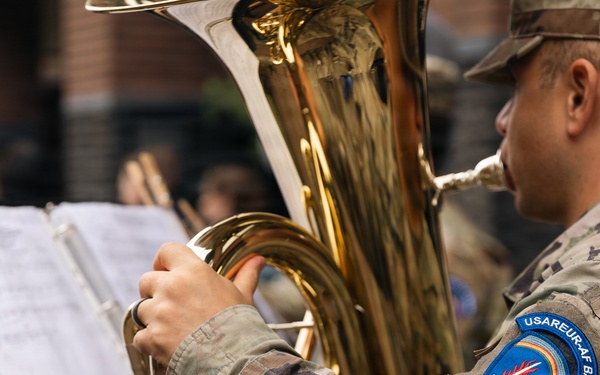 A U.S. Army soldier assigned to the USAREUR-AF Band is playing a tuba during what appears to be an outdoor musical performance. The soldier is wearing MultiCam (OCP) camouflage uniform with a visible unit patch on the left shoulder reading 'USAREUR-AF BAND'. Sheet music on a stand is visible in the foreground, indicating a structured, rehearsed performance.