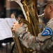 A U.S. Army soldier assigned to the USAREUR-AF Band is playing a tuba during what appears to be an outdoor musical performance. The soldier is wearing MultiCam (OCP) camouflage uniform with a visible unit patch on the left shoulder reading 'USAREUR-AF BAND'. Sheet music on a stand is visible in the foreground, indicating a structured, rehearsed performance.