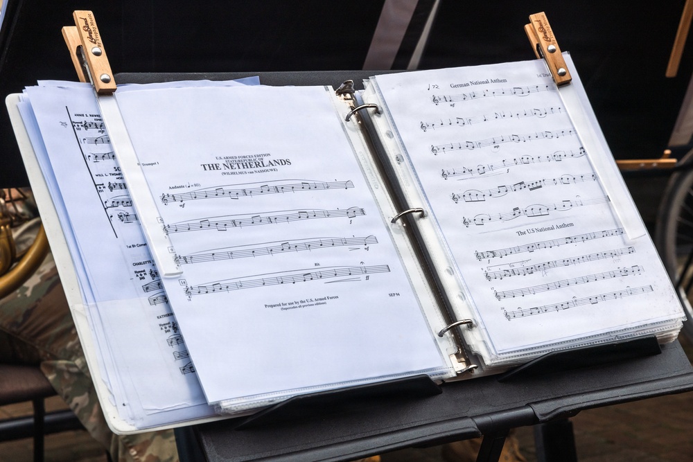 A music stand holds an open binder containing sheet music prepared for U.S. Armed Forces use. The binder displays multiple pieces including the State Anthem of The Netherlands (Wilhelmus van Nassouwe), the German National Anthem, and the US National Anthem. Two wooden clothespins are clipped to the top of the binder to secure the pages against wind.
