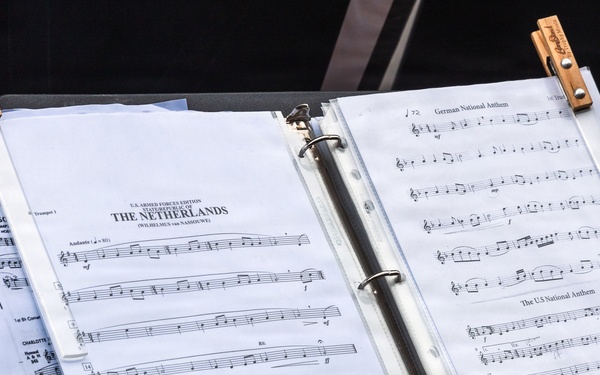 A music stand holds an open binder containing sheet music prepared for U.S. Armed Forces use. The binder displays multiple pieces including the State Anthem of The Netherlands (Wilhelmus van Nassouwe), the German National Anthem, and the US National Anthem. Two wooden clothespins are clipped to the top of the binder to secure the pages against wind.