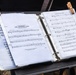 A music stand holds an open binder containing sheet music prepared for U.S. Armed Forces use. The binder displays multiple pieces including the State Anthem of The Netherlands (Wilhelmus van Nassouwe), the German National Anthem, and the US National Anthem. Two wooden clothespins are clipped to the top of the binder to secure the pages against wind.