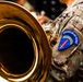 A close-up photograph of a military musician in U.S. Army camouflage (OCP pattern) uniform holding a large brass instrument, likely a tuba or euphonium. The soldier's shoulder patch clearly identifies them as a member of the USAREUR-AF Band (United States Army Europe and Africa Band). The image emphasizes both the gleaming brass bell of the instrument and the distinctive unit patch.