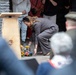 A woman in a gray suit is bending down to place a floral wreath or bouquet at the base of a wooden podium during what appears to be a formal commemorative ceremony. The podium bears an engraved emblem with the partial text 'ALIS' visible. An audience is gathered around, suggesting this is a public memorial or dedication event.