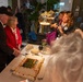 A formal indoor gathering or celebration is taking place around a decorated sheet cake on a table. An elderly man wearing a black cap and a woman in a red jacket stand near the cake, while a photographer with a camera and lanyard documents the moment. Military personnel in camouflage uniforms are visible in the background.