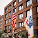 A U.S. Army color guard of four soldiers in MultiCam (OCP) uniforms and black berets stands at attention on a wet brick plaza in front of a modern red-brick building. They are presenting the U.S. national flag and what appears to be a U.S. Army organizational flag with gold fringe, while two of the soldiers carry ceremonial rifles. The setting and architecture suggest a location in Western Europe, likely the Netherlands or Belgium.