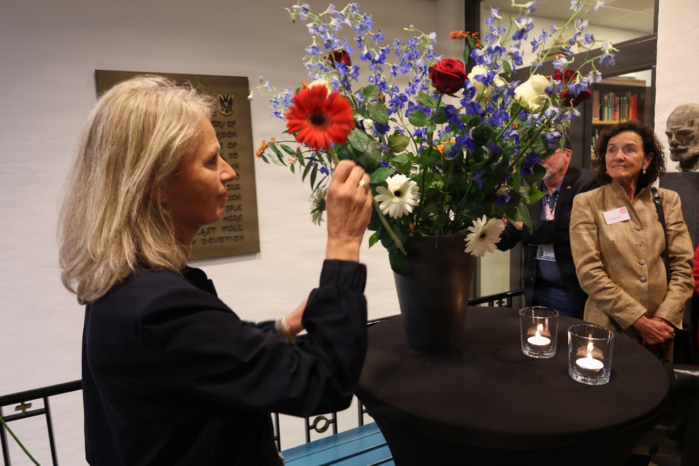 A woman with gray-blonde hair arranges a large floral bouquet containing red gerberas, blue delphiniums, red roses, and white daisies in a dark vase on a round table. Two lit tea candles are placed on the same table. A second woman in a tan blazer with a name badge observes the arrangement, while a man stands in the background near a bookshelf.