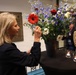 A woman with gray-blonde hair arranges a large floral bouquet containing red gerberas, blue delphiniums, red roses, and white daisies in a dark vase on a round table. Two lit tea candles are placed on the same table. A second woman in a tan blazer with a name badge observes the arrangement, while a man stands in the background near a bookshelf.