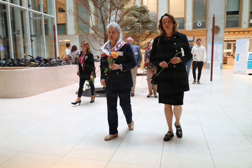 Two women dressed in dark formal attire walk together through what appears to be a modern institutional building atrium, each carrying single flowers. The foreground woman wears a patriotic red, white, and blue scarf and holds an orange flower, while the woman beside her holds a white flower. Several other people are visible in the background of the well-lit interior space.