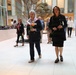 Two women dressed in dark formal attire walk together through what appears to be a modern institutional building atrium, each carrying single flowers. The foreground woman wears a patriotic red, white, and blue scarf and holds an orange flower, while the woman beside her holds a white flower. Several other people are visible in the background of the well-lit interior space.
