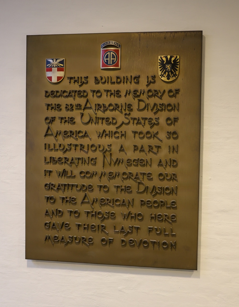 A bronze commemorative plaque mounted on a white wall, dedicated to the 82nd Airborne Division of the United States Army for their role in liberating Nijmegen during World War II. The plaque features three military/civic insignia at the top and raised lettering expressing gratitude to the division and the American people. It honors those who gave their lives during the liberation.