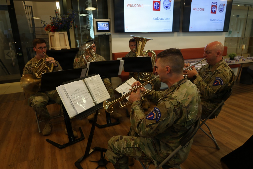 Five U.S. Army soldiers in camouflage uniforms perform as a brass ensemble in what appears to be a hospital or institutional lobby. The welcome screens in the background display the 82nd Airborne Division patch and 'Radboudumc' branding, suggesting this is a performance at a Dutch medical center. The musicians are playing trumpet, French horn, and tuba/euphonium instruments while reading from sheet music on stands.