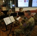 Five U.S. Army soldiers in camouflage uniforms perform as a brass ensemble in what appears to be a hospital or institutional lobby. The welcome screens in the background display the 82nd Airborne Division patch and 'Radboudumc' branding, suggesting this is a performance at a Dutch medical center. The musicians are playing trumpet, French horn, and tuba/euphonium instruments while reading from sheet music on stands.