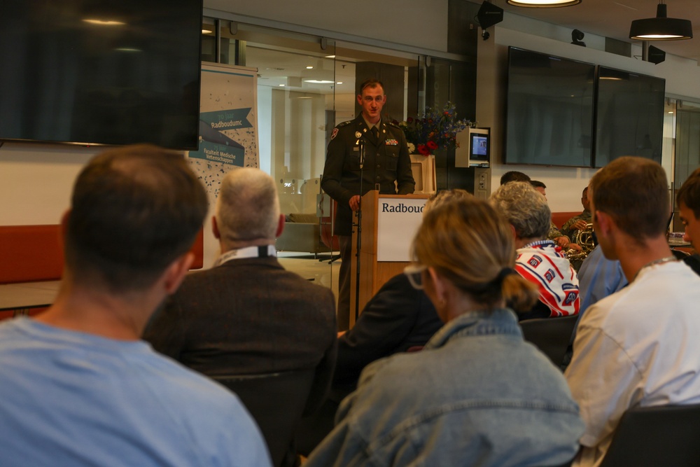 A military officer in dress uniform is delivering a speech at a podium bearing the 'Radboudumc' logo, likely at Radboud University Medical Center in the Netherlands. An audience of civilians and at least one other military personnel are seated facing the speaker. The setting appears to be a modern conference or lecture room.